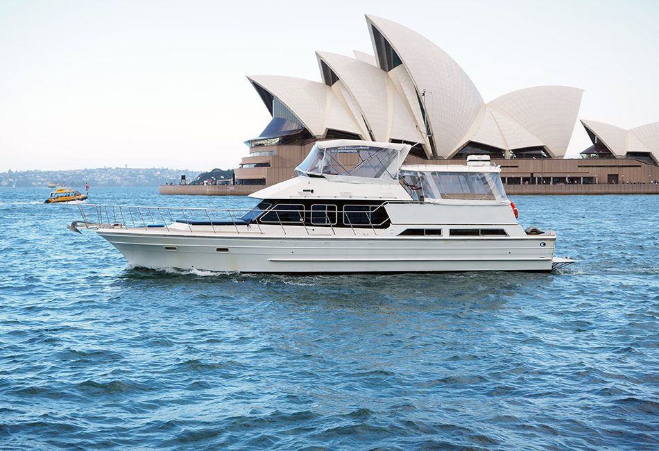 A white yacht from The Yacht Social Club sails near the iconic Sydney Opera House with its distinctive sail-like design structures. A small ferry is visible in the background on the blue waters of Sydney Harbour. The clear sky enhances this scenic view, perfect for Boat Parties by The Yacht Social Club.