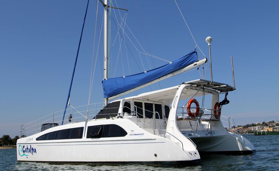 A white catamaran sailboat with a blue sail cover sits on the water against a backdrop of buildings. The boat has "Catalpa" written on the side and two red life rings at the back. The sky is clear, and the water is calm, perfect for an event with The Yacht Social Club Sydney Boat Hire.