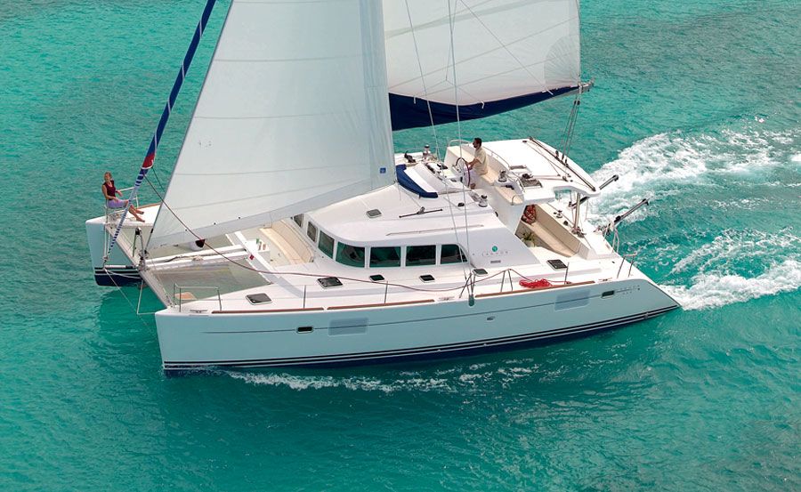 A white sailboat with two large sails glides through clear, turquoise waters. Two people are visible on deck, enjoying the sunny day and the ocean surroundings. The boat creates a small wake as it moves forward, perfect for an exclusive outing like The Yacht Social Club event.