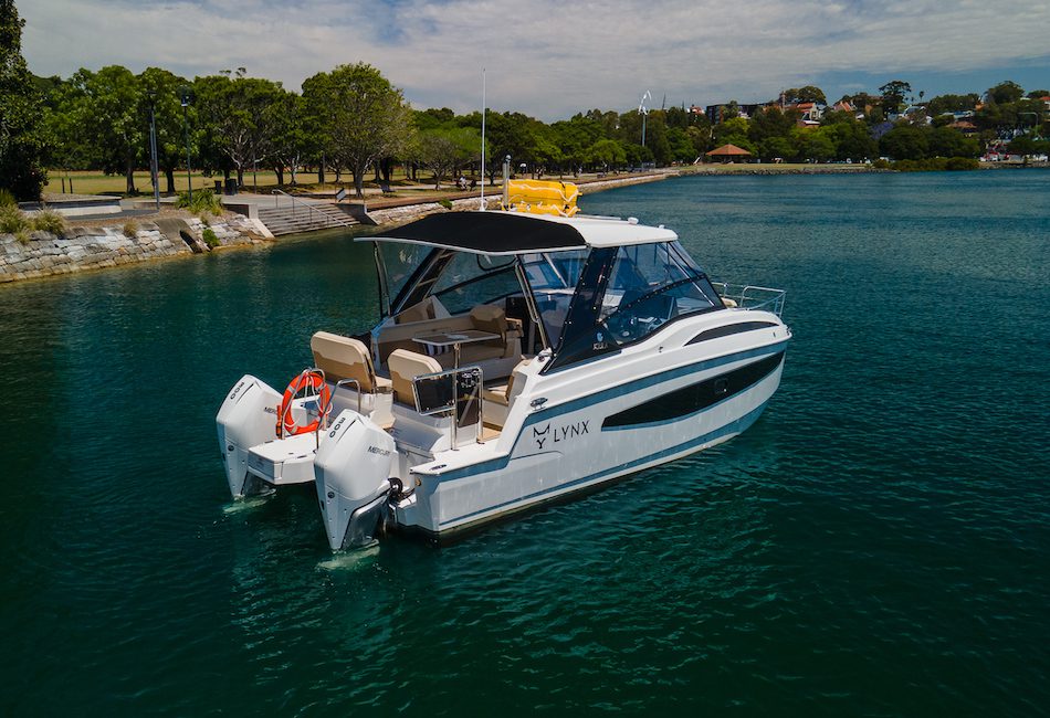 A sleek white motorboat named "LYNX" floats on calm waters near a tree-lined shore. The boat features a black rooftop canopy and two outboard motors. The clear sky and distant houses add to the serene atmosphere. A life buoy is visible on the back deck, making it perfect for Boat Parties with The Yacht Social Club Sydney Boat Hire.