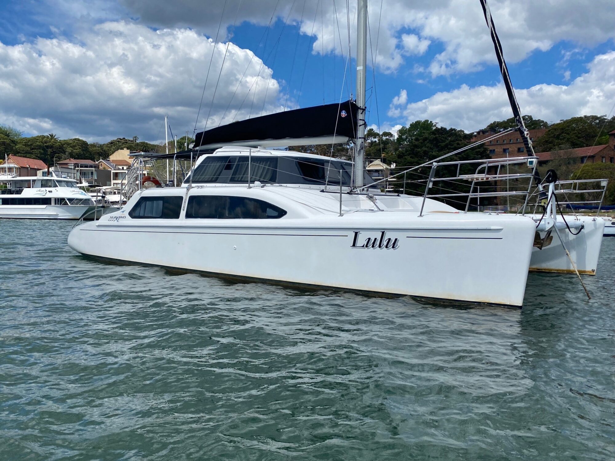 A white catamaran sailboat named "Lulu" is docked on calm waters. The vessel features a sleek design with large windows and a black mast. The backdrop includes other boats, trees, and buildings under a partly cloudy sky—ideal for Boat Parties Sydney with The Yacht Social Club.