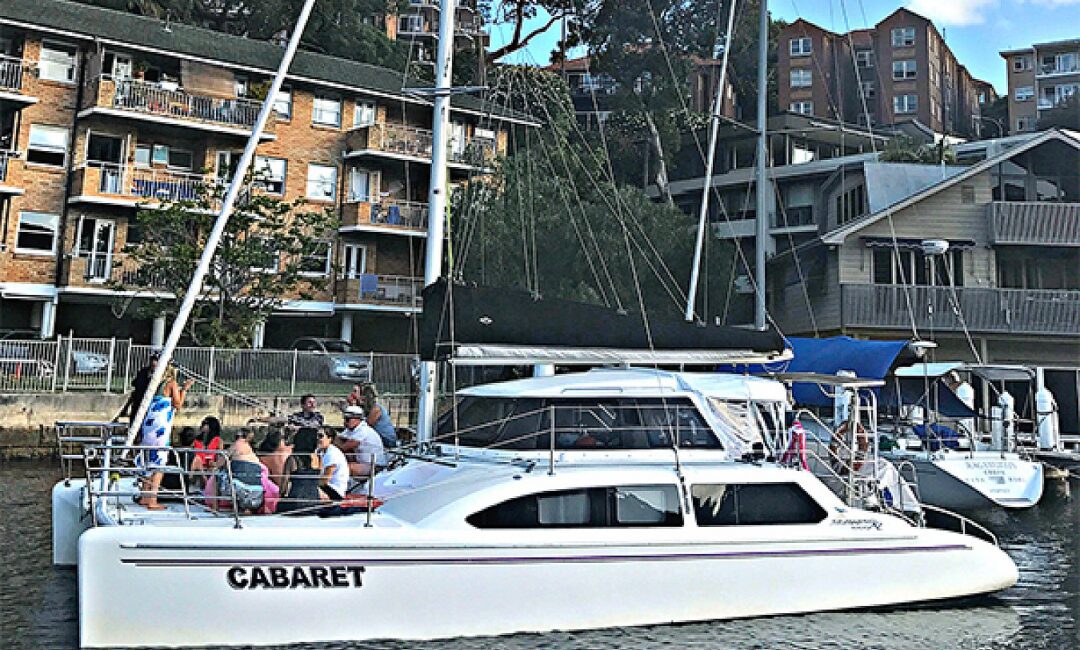 A group of people sit on a white boat named "Cabaret," docked alongside a marina with apartment buildings in the background. The boat, perfect for Boat Parties Sydney by The Yacht Social Club, features a sail and several windows. The water appears calm with surrounding greenery and multiple boats.