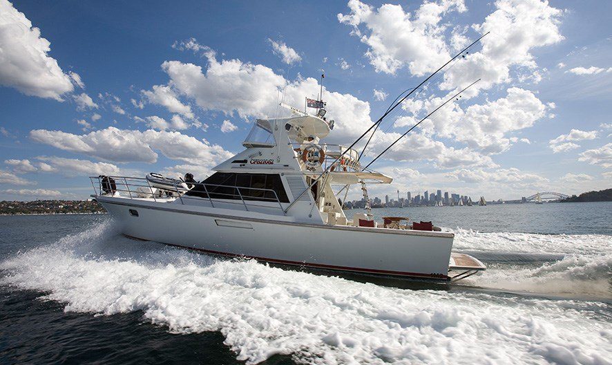 A white yacht with fishing rods on its deck cruises through the water, creating waves behind it. The sky is filled with scattered clouds, and a city skyline is visible in the background. Experience moments like these with Boat Parties Sydney by The Yacht Social Club.