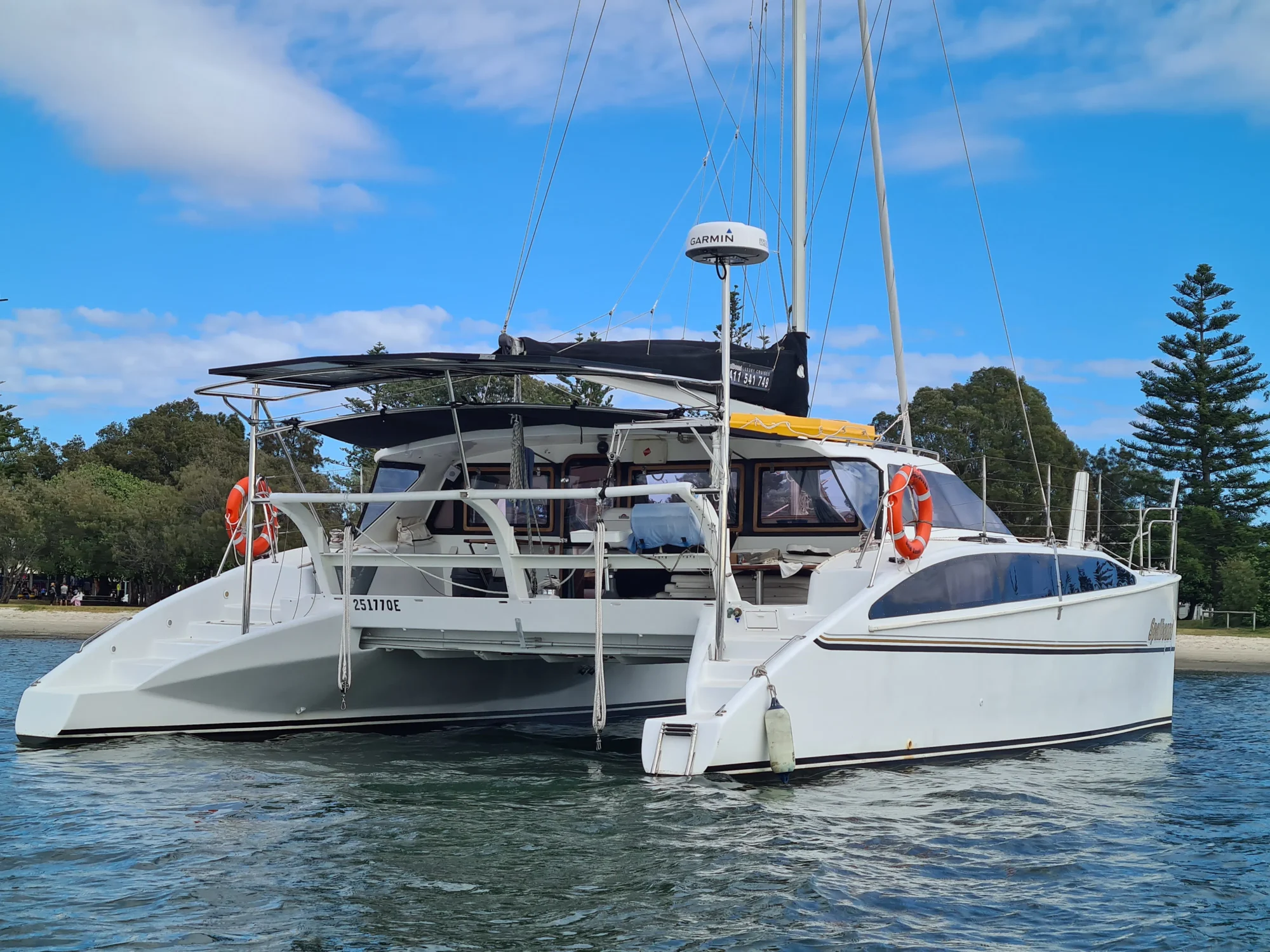 A modern catamaran anchored in calm waters near a shoreline features white hulls, solar panels on the rooftop, life rafts, and an array of navigation equipment. Lush greenery and tall pine trees are visible in the background under a partly cloudy sky. Perfect for luxury yacht rentals Sydney.