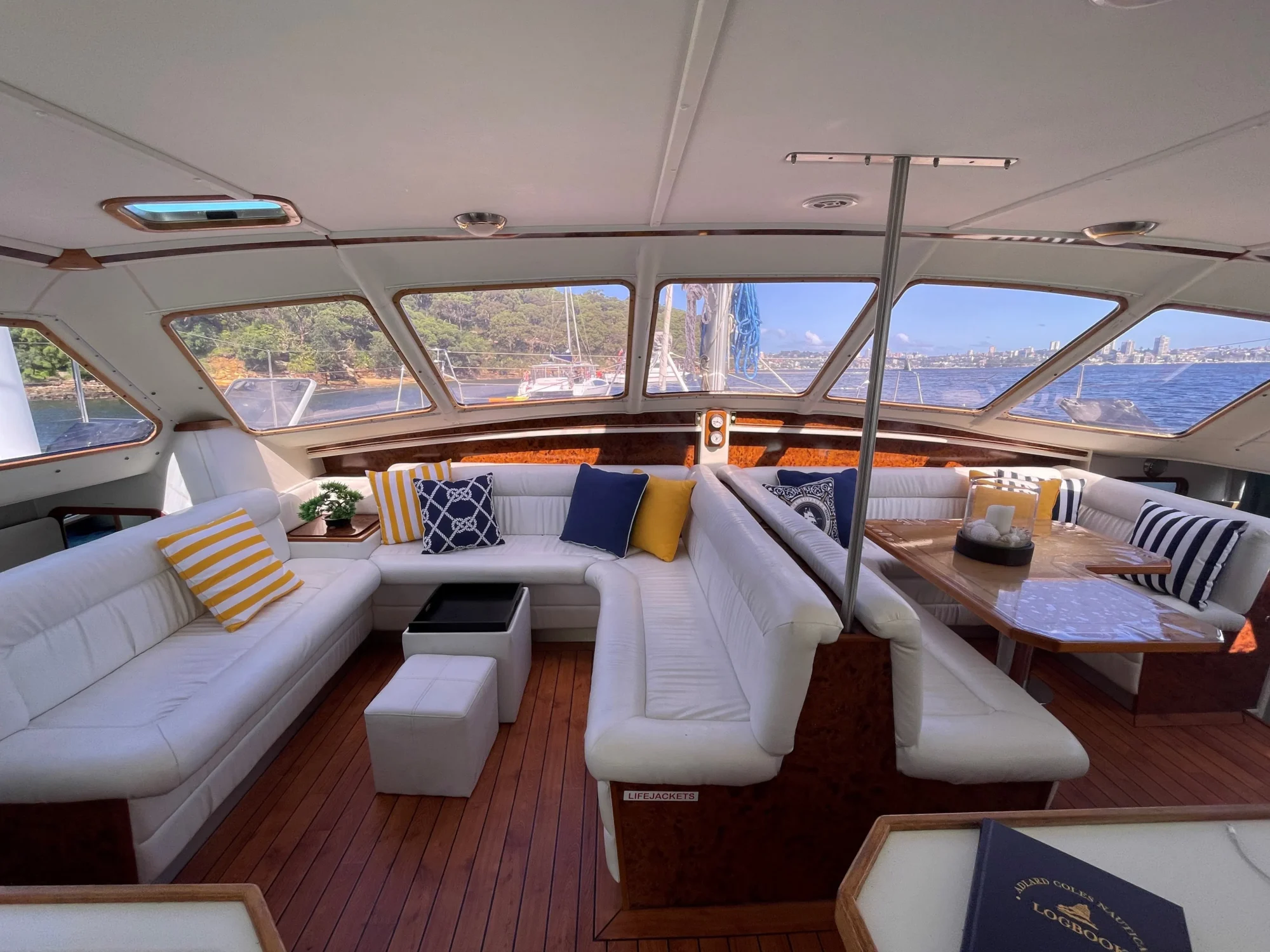 A luxurious yacht interior featuring white leather seating with colorful throw pillows, a wooden floor, and large windows providing panoramic views of the sea and distant cityscape. A small table with decorative items is also visible near the windows. Perfect for Boat Rentals and Parties in Sydney by The Yacht Social Club.