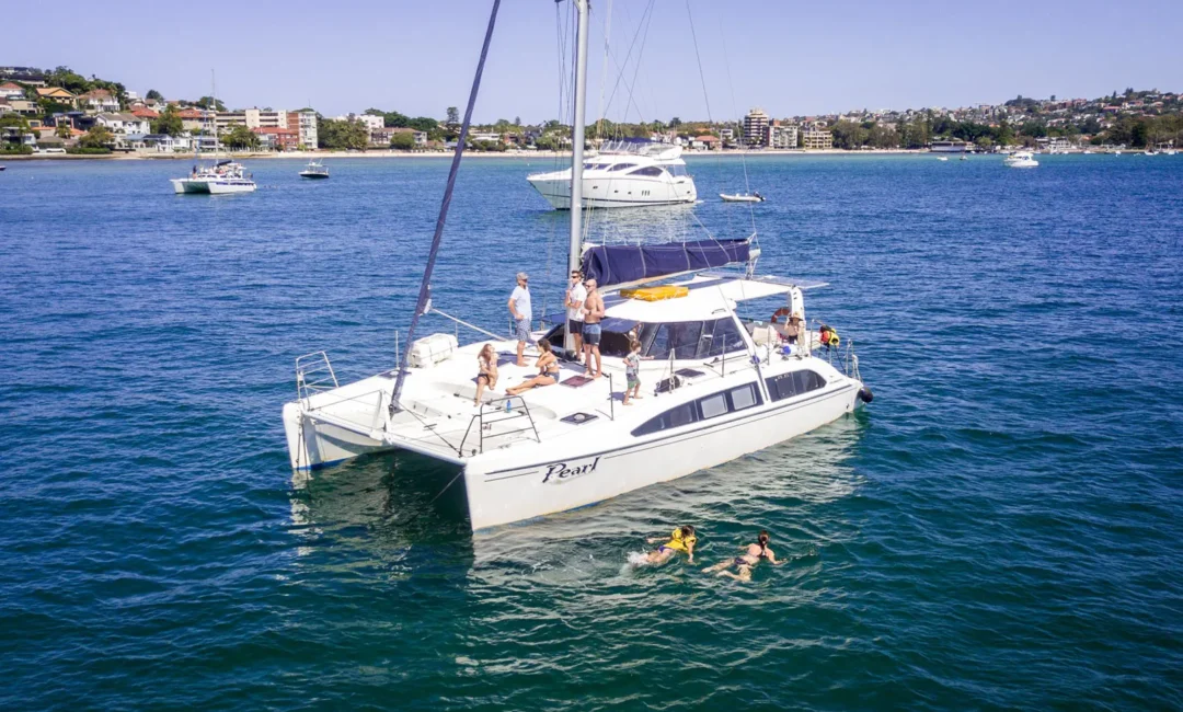 A catamaran named 'Pearl' floats on calm blue waters near a coastal town. Several people are on board, relaxing and enjoying the sun, while a few others swim nearby. The Yacht Social Club Event Boat Charters offer similar experiences, with other boats dotted across the water and residential buildings visible in the background.