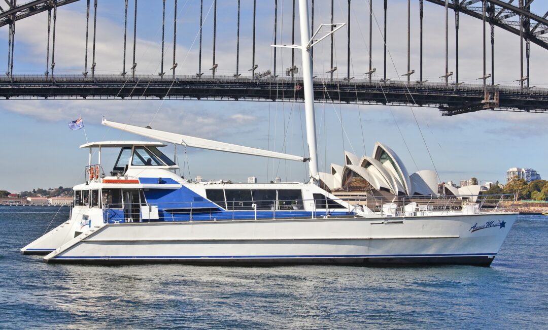 A white and blue catamaran sails on a sunny day, with the iconic Sydney Harbour Bridge and Sydney Opera House in the background. The boat, part of The Yacht Social Club, has a flag and several windows along its side. The water is calm and the sky partly cloudy.