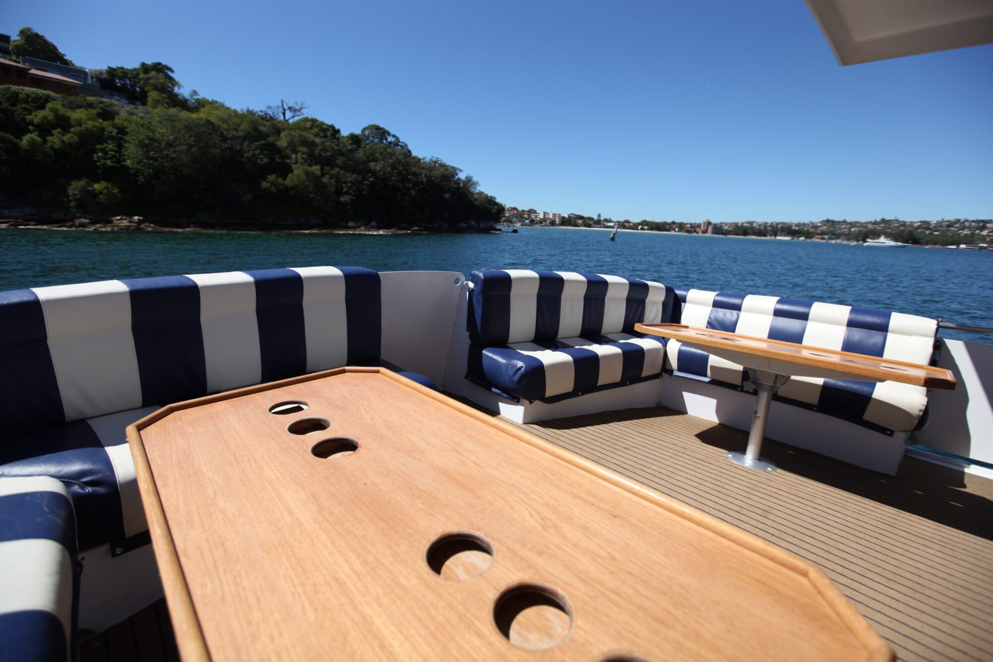 The image shows the deck of a boat with wooden tables and cushioned seating. The seats feature blue and white striped patterns. In the background, calm water, a green treeline, and distant shoreline buildings are visible under a clear blue sky – perfect for The Yacht Social Club Event Boat Charters.