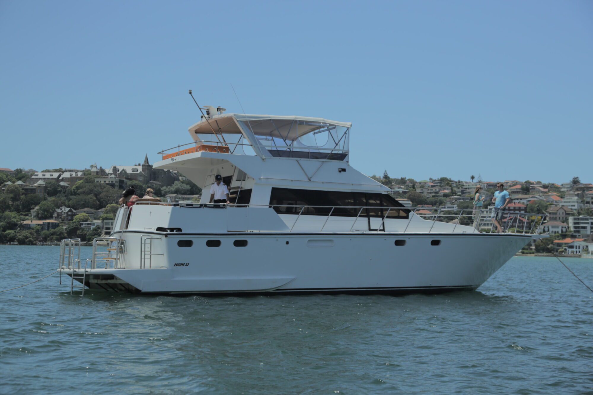 A white yacht with multiple decks is anchored on a calm water body. A few people are enjoying themselves on the boat, and in the background, a residential area with houses on a hillside is visible under a clear blue sky—perfect for Boat Parties Sydney The Yacht Social Club.