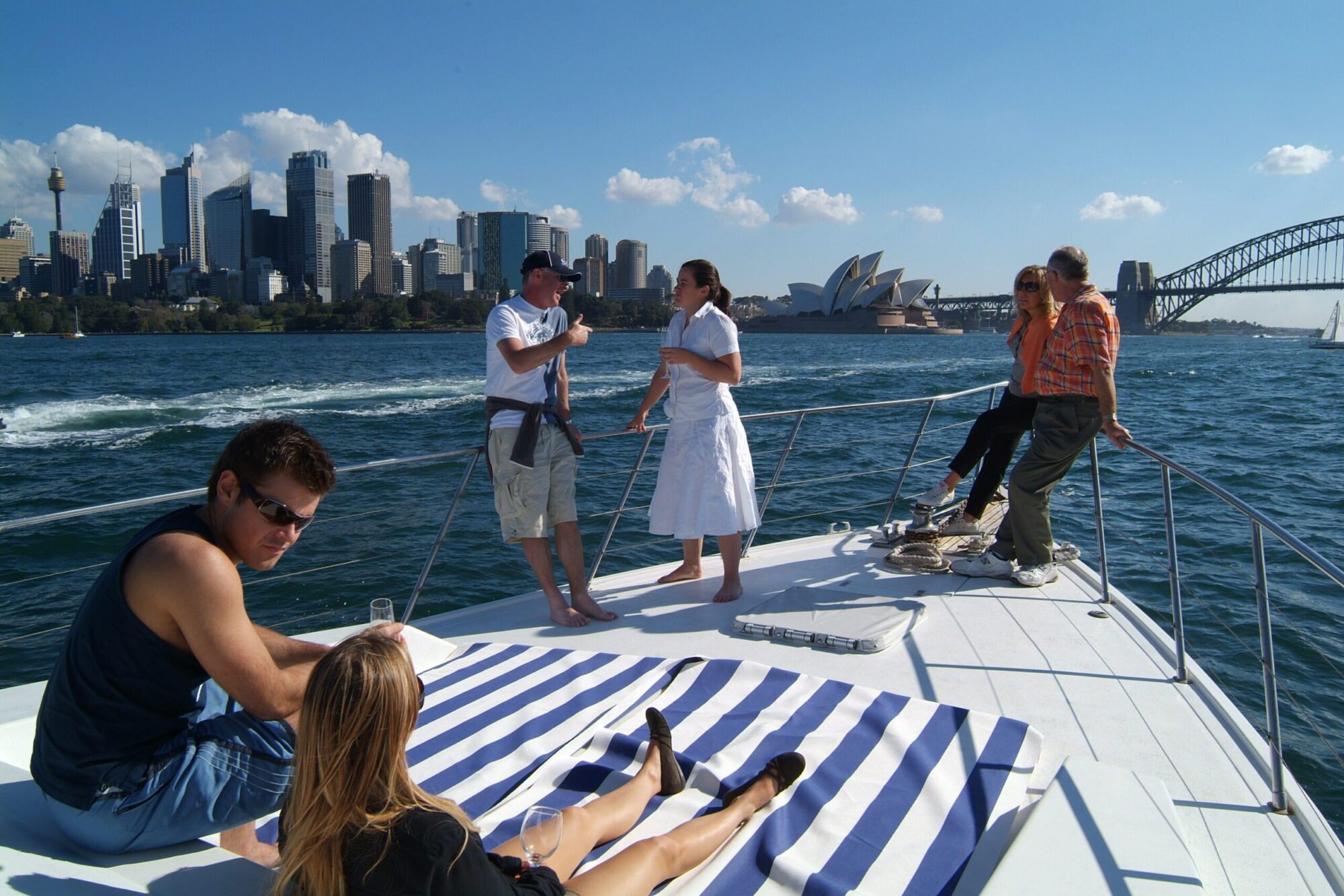 A group of people enjoy a sunny day on a boat rental and parties with The Yacht Social Club. Two men and a woman stand talking near the front, while another man and woman sit at the edge. In the foreground, a man and a woman relax on a striped towel. Sydney Opera House and Sydney Harbour Bridge are visible.