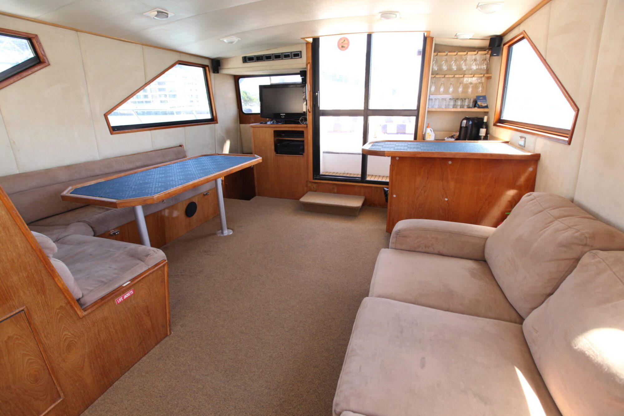 A cozy boat interior featuring a beige couch on the right, a wood-paneled bar area with a coffee maker and glasses in the back, a TV on a wooden stand, and a blue-topped dining table with built-in seating on the left. Windows offer natural light and outside views, perfect for boat parties by The Yacht Social Club Sydney Boat Hire.