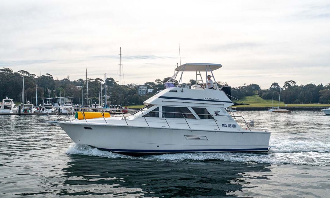 A white motor yacht named "Monty Fuel" is cruising through a calm waterway. The yacht features a cabin with windows and an upper deck with seating, perfect for boat parties organized by The Yacht Social Club Sydney Boat Hire. Background includes other docked boats, trees, and a grassy area under an overcast sky.