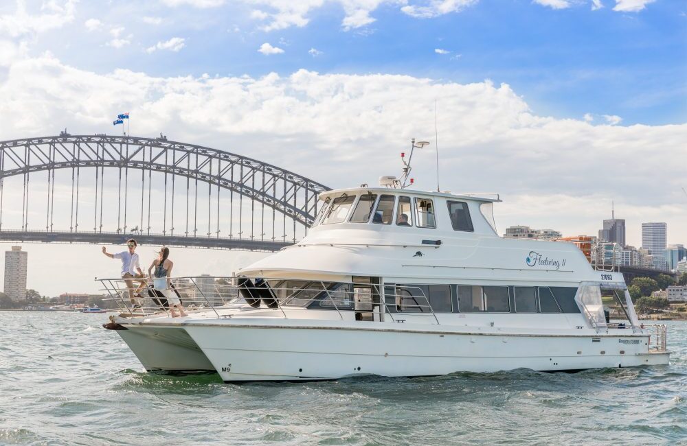 A white yacht sails on a sunny day with two people standing on its deck, enjoying the view. The iconic Sydney Harbour Bridge is visible in the background, along with a partly cloudy sky and scattered city buildings. Experience this moment with Luxury Yacht Rentals Sydney for an unforgettable adventure.