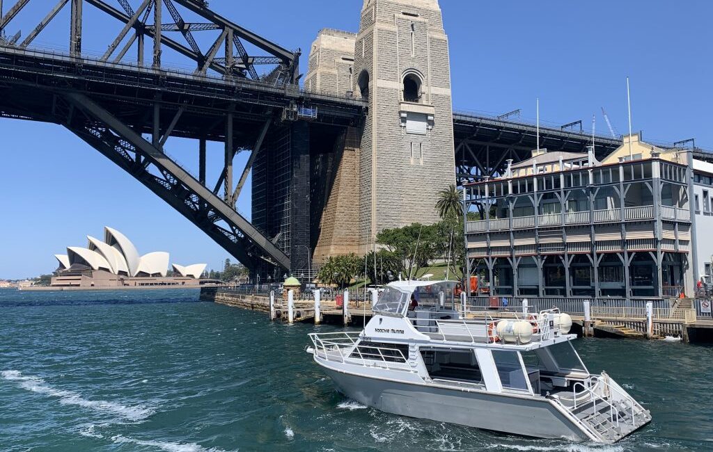 A boat from The Yacht Social Club floats on blue water near a pier with a large bridge overhead. The Sydney Opera House can be seen in the distance under a clear sky. The bridge has a tall stone pillar, and old-style buildings stand near the waterfront, enhancing the charm of Sydney Harbour.