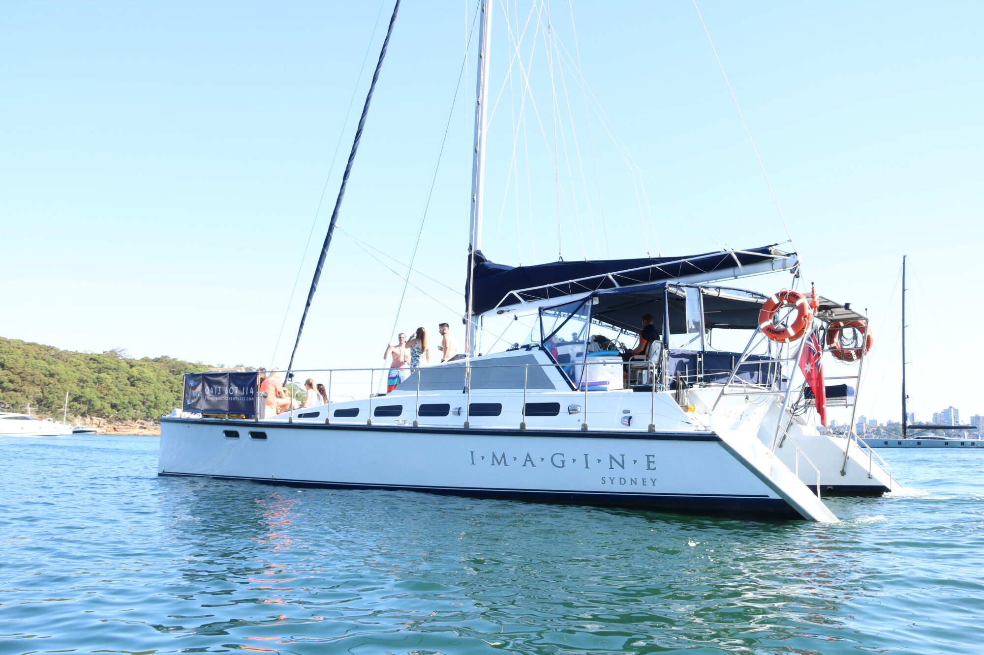 A white sailboat named "IMAGINE SYDNEY" is floating on a calm body of water. The boat, available for Sydney Harbour Boat Hire The Yacht Social Club, has multiple people on board and a forested shore in the background under a clear blue sky. Two red life rings are visible on the side of the boat.