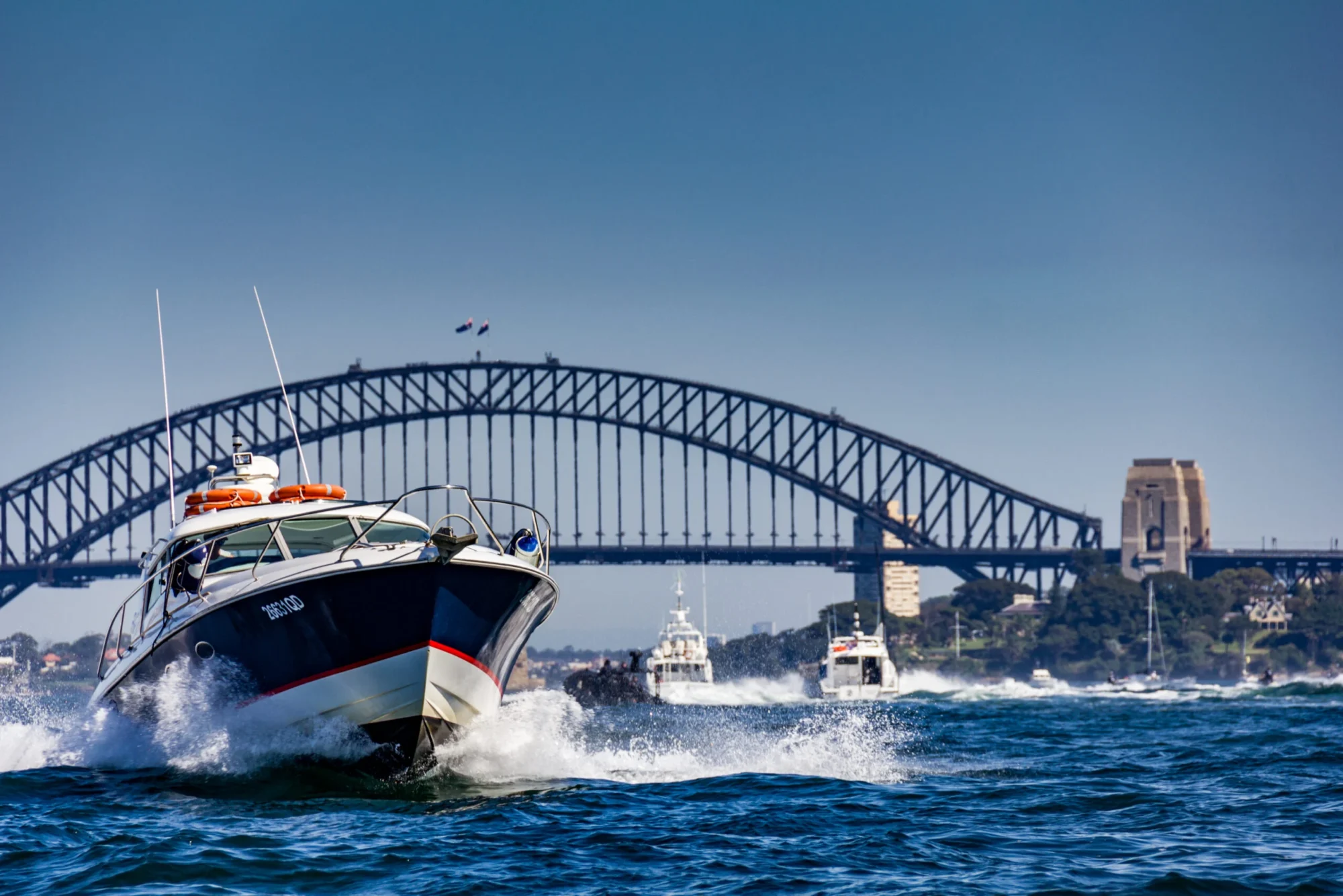 A speedboat creates white waves as it moves swiftly across the water with the Sydney Harbour Bridge in the background. Other boats and yachts, part of The Yacht Social Club Sydney Boat Hire, are visible behind it in the blue water, and the sky is clear and bright.