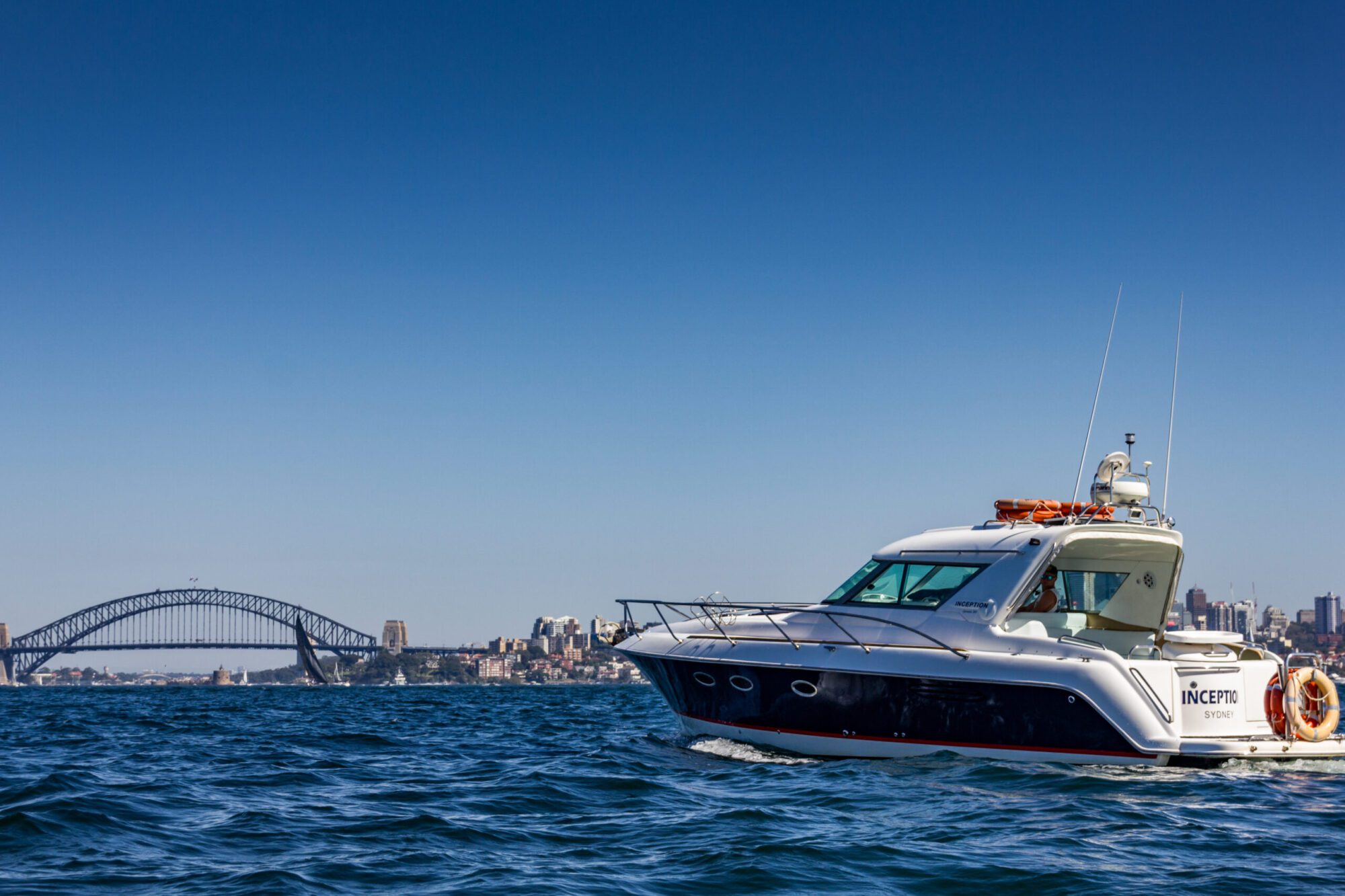 A motor yacht named "Inception" cruises on a body of water with a cityscape in the background, featuring the iconic Sydney Harbour Bridge under a clear blue sky. Ideal for The Yacht Social Club Event Boat Charters, it's perfect for experiencing luxury on Sydney Harbour.