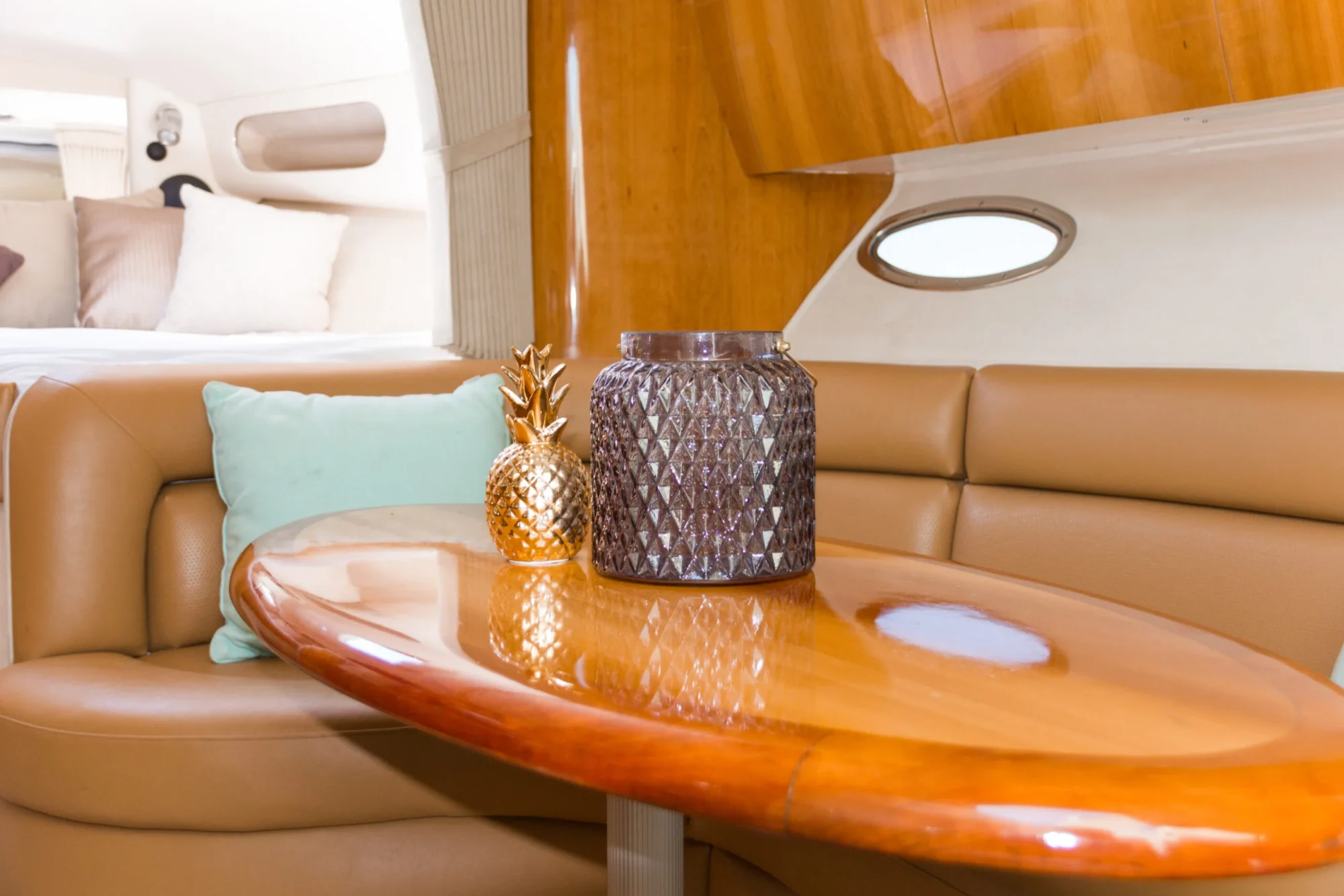 Interior of a luxury boat featuring a small wooden table with a gold pineapple ornament and a dark patterned vase in the center. The seating consists of plush tan leather benches with light blue pillows. Rounded window and cabin details are visible in the background, perfect for Boat Parties Sydney The Yacht Social Club.