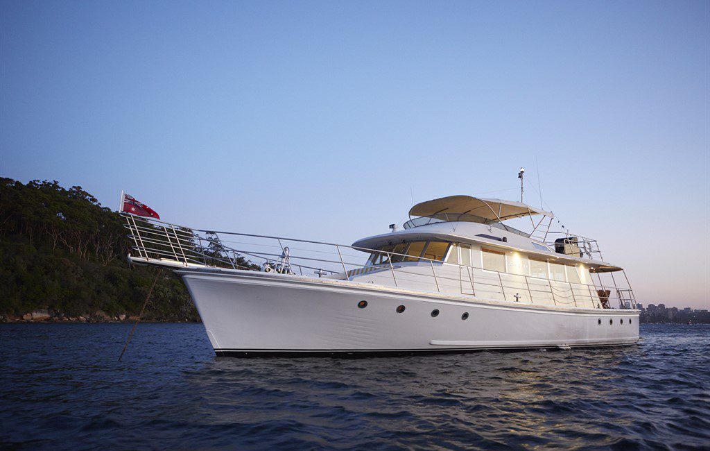 A luxurious white yacht with multiple decks is moored on calm waters during twilight. The vessel, perfect for Boat Parties Sydney by The Yacht Social Club, features railings, a canopy on the upper deck, and a red flag at the stern. Trees and a rocky shoreline are visible in the background under a clear sky.