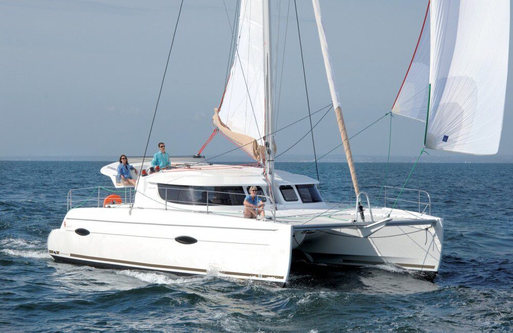 A white catamaran with multiple sails is sailing on a calm sea. Three people are onboard, enjoying the ride; one is sitting at the front, another is standing near the mast, and the third is at the back near the helm. The sky above is clear and blue—a perfect day for Sydney Harbour Boat Hire with The Yacht Social Club.