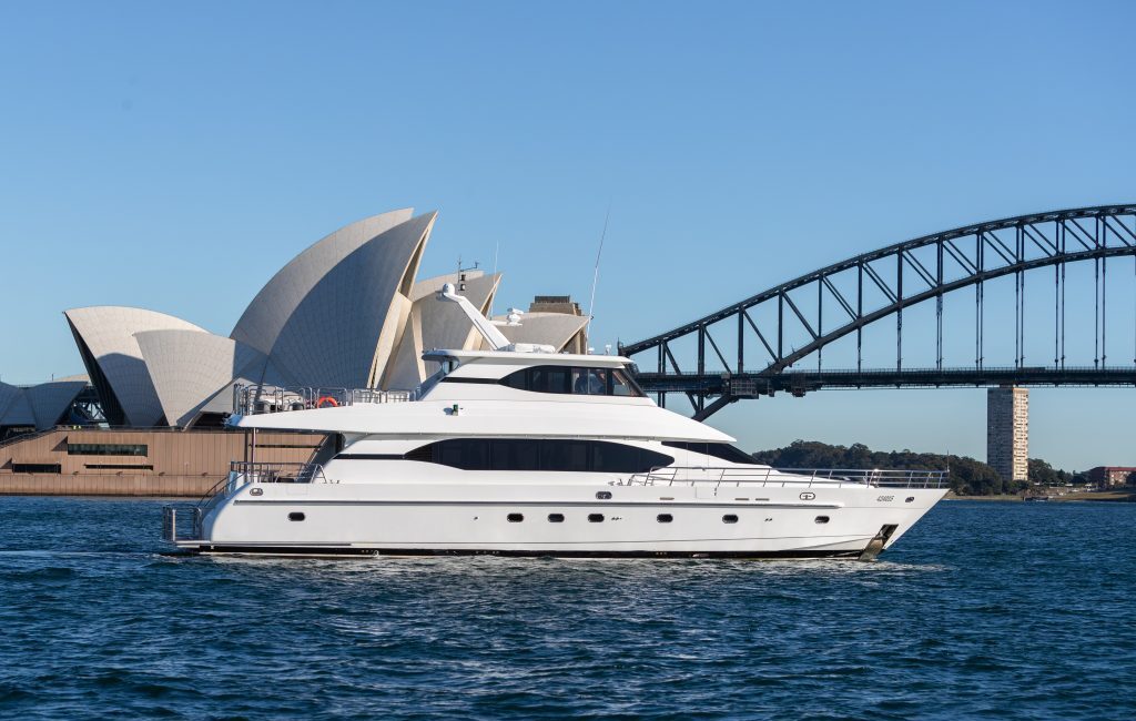 A white yacht cruises through the water with the Sydney Opera House and Sydney Harbour Bridge in the background on a clear, sunny day, exemplifying The Yacht Social Club's premium Sydney boat hire experience.