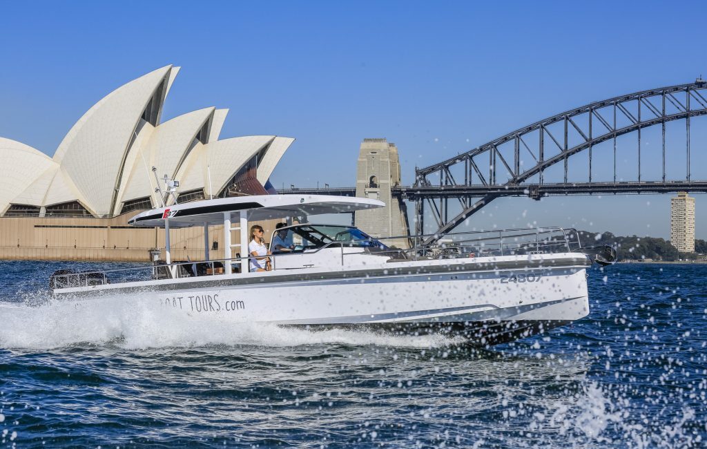 A motorboat from "Boating Tours" speeds through the water in front of the Sydney Opera House and Sydney Harbour Bridge under a clear blue sky. Fine splashes of water are seen as it moves across the scene, perfect for those considering Boat Parties Sydney The Yacht Social Club.