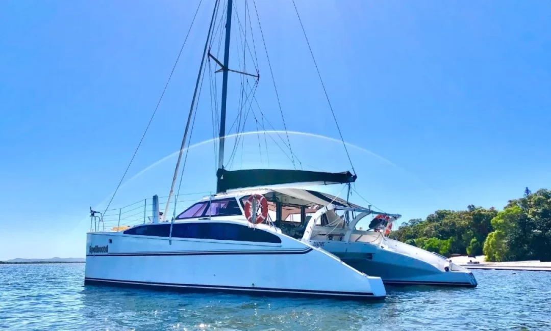 A white catamaran is anchored in clear blue waters. The boat, hired from The Yacht Social Club Sydney Boat Hire, has a green canopy and multiple masts. People are seen relaxing under the canopy. The background includes green, tree-covered land under a cloudless, sunny sky.