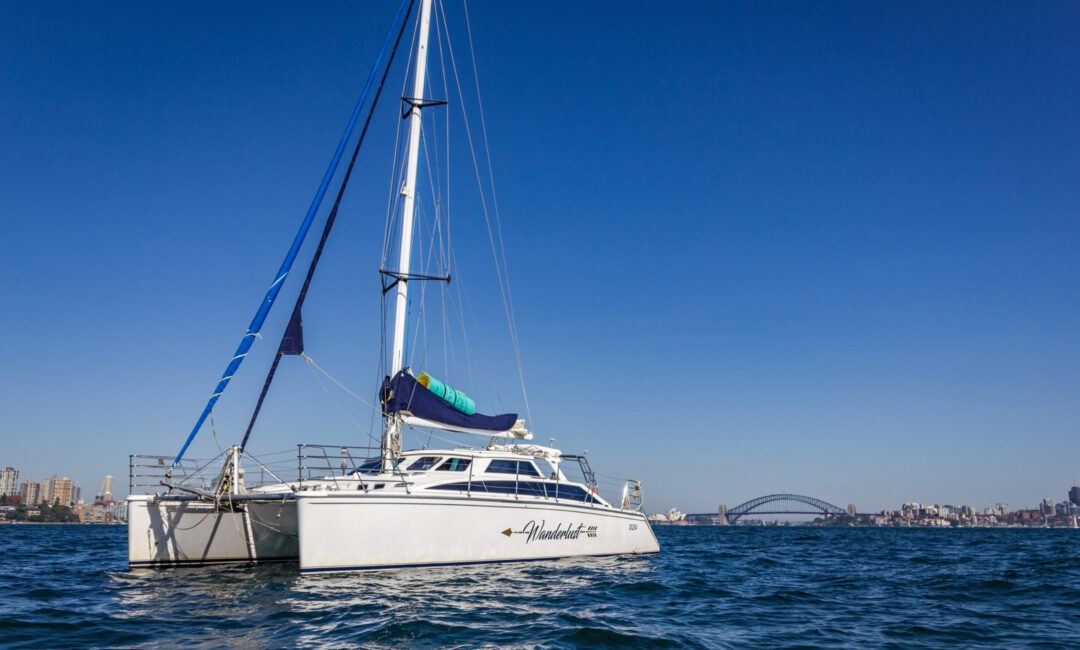 A white catamaran sails on calm blue water with a skyline and a distinct arch bridge in the background under a clear blue sky. The boat's sail is partially visible, featuring a blue and green rolled-up sailcloth. The cityscape, part of The Yacht Social Club Sydney Boat Hire, features several buildings.