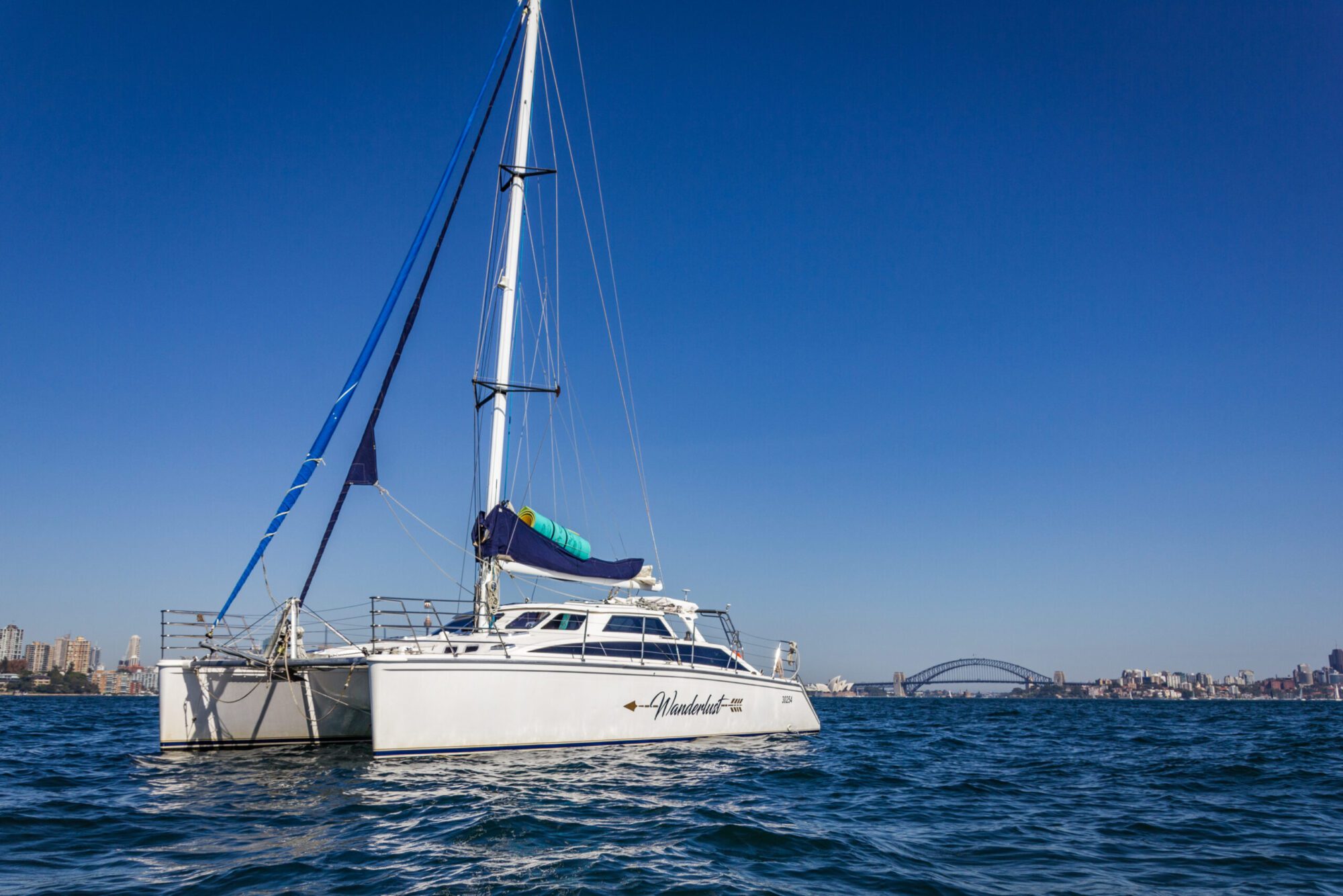 A white catamaran sails on calm blue water with a skyline and a distinct arch bridge in the background under a clear blue sky. The boat's sail is partially visible, featuring a blue and green rolled-up sailcloth. The cityscape, part of The Yacht Social Club Sydney Boat Hire, features several buildings.