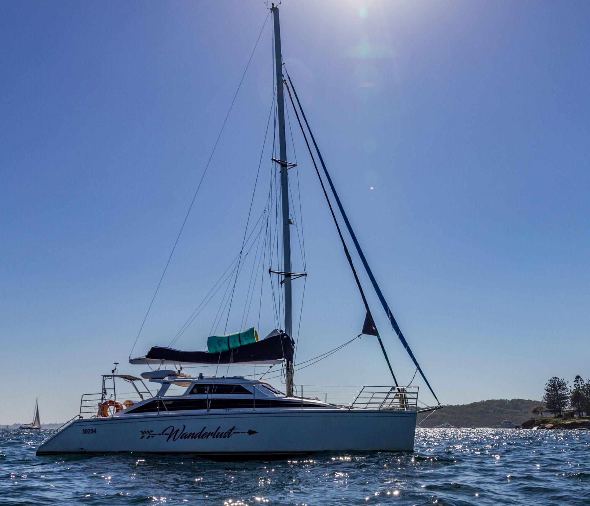 A white catamaran named "Wanderlust" sails on a blue, sunlit sea. The vessel has a blue sail and a deck with visible railings. Calm waters and bright sunshine surround the boat, with distant land in the background—an idyllic scene perfect for The Yacht Social Club Sydney Boat Hire.