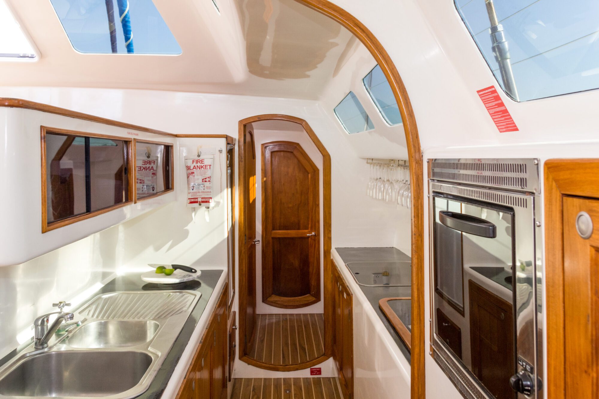 The interior of a sailboat features a compact kitchen with a stainless steel sink, stove, and oven. Wooden cabinets and several skylights brighten the space. Wine glasses hang above the counter, adding an elegant touch. Perfect for *Luxury Yacht Rentals Sydney* or *Boat Parties Sydney The Yacht Social Club*.