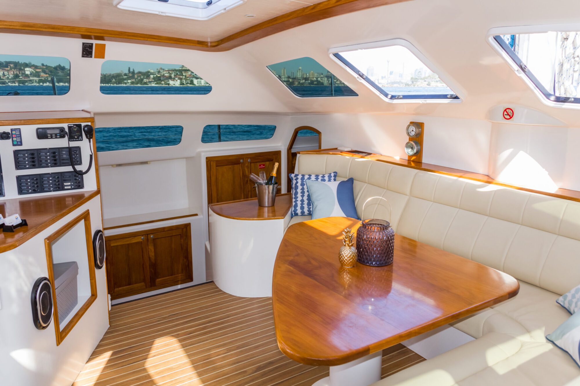 Interior of a luxury yacht with comfortable seating, a wooden dining table, and nautical decorations. Large windows offer stunning views of the ocean and shoreline, while natural light floods the space. Perfect for Sydney Harbour Boat Hire The Yacht Social Club gatherings, it features built-in cupboards and control panels.