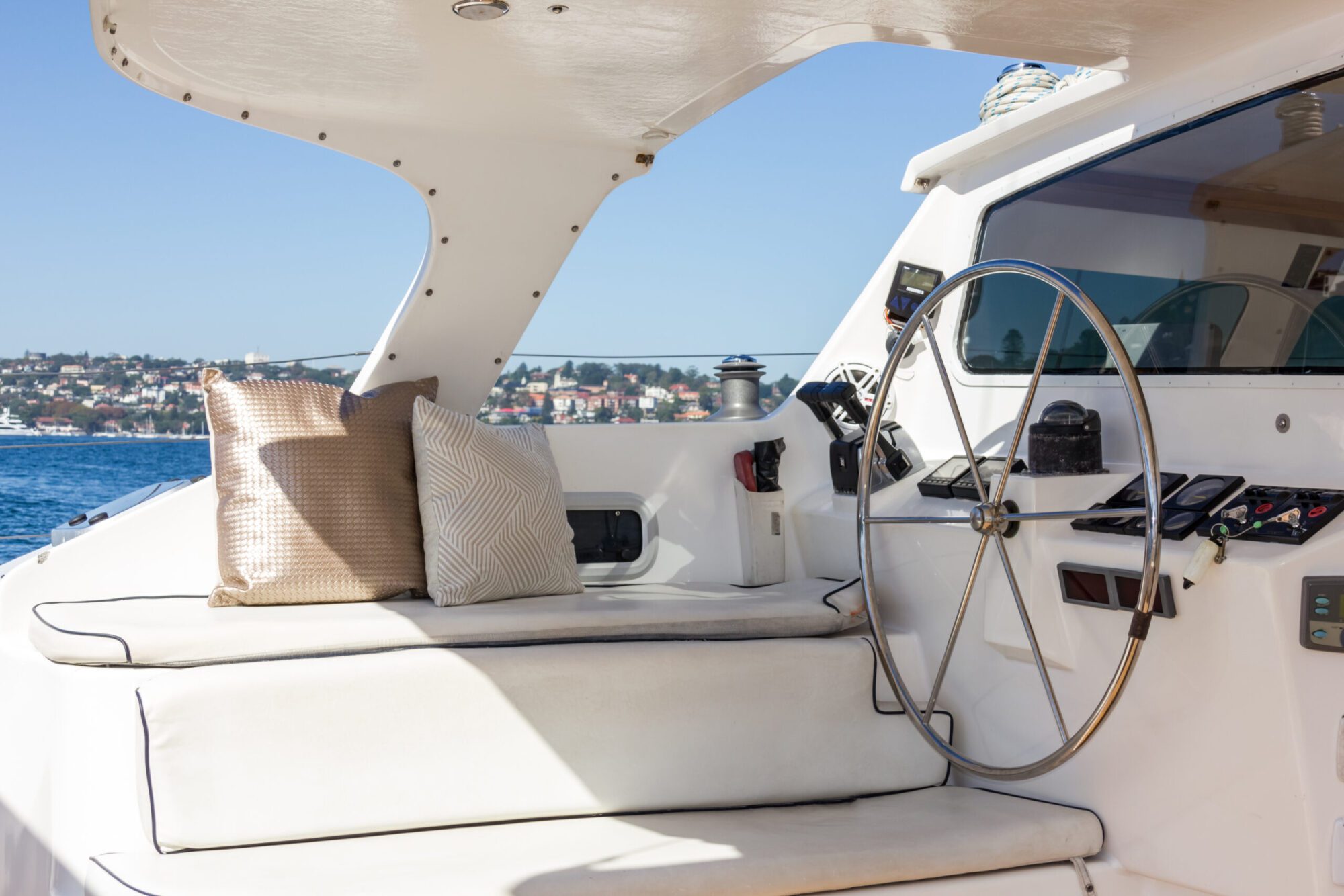 A close-up of a yacht's helm featuring a large steering wheel, various navigational instruments, and a cushioned bench seat with two decorative pillows. The yacht is on clear blue water with a coastal town visible in the background, perfect for *The Yacht Social Club Event Boat Charters* experience.