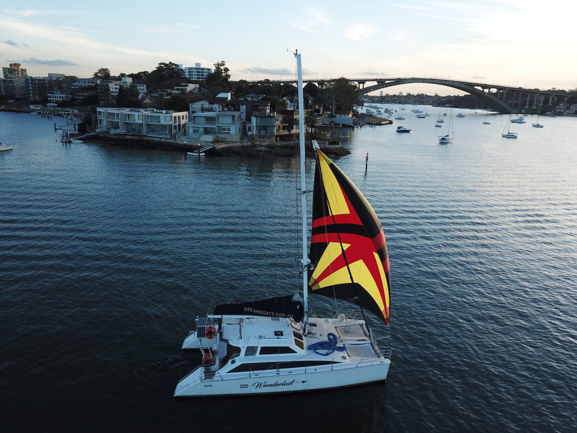 A white catamaran with colorful sails, featuring a striking yellow, red, and black design, glides on calm waters. Distant houses, boats, and a large arched bridge are visible in the background under a clear blue sky. Perfect for Sydney Harbour boat hire by The Yacht Social Club.