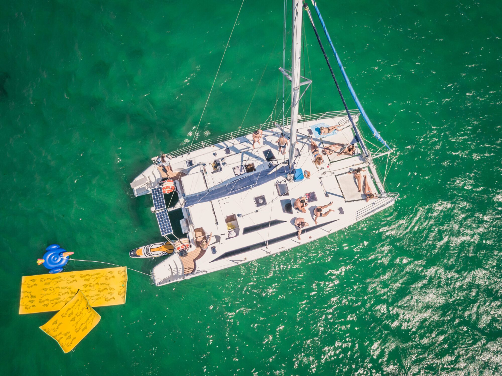 Aerial view of a white catamaran with people lounging on its deck, anchored in clear green water. A blue inflatable tube is tied to the boat, and two yellow floats are placed beside it in the water, providing a relaxed, tropical setting. Experience this with The Yacht Social Club’s exclusive Boat Parties in Sydney.