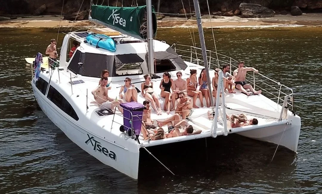 A group of people relax on a white catamaran named "X-Sea" anchored in calm water near a rugged shoreline. Some lounge on the deck, while others sit or stand, enjoying their time under the sun. Various items, including a kayak and bags, are on the boat. Perfect for Boat Rental and Parties Sydney The Yacht Social Club.