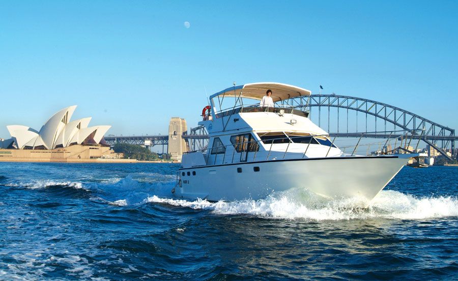 A luxury yacht cruises on a sunny day with the Sydney Opera House and the Sydney Harbour Bridge visible in the background, set against a clear blue sky and calm waters, offering an idyllic scene perfect for Yacht Rentals by The Yacht Social Club.