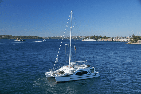 A white multi-hull yacht from The Yacht Social Club sails on a bright blue body of water under a clear, sunny sky. In the background, another ship is visible near the coastline and a distant shoreline with buildings and trees can be seen. The scene is serene and picturesque.