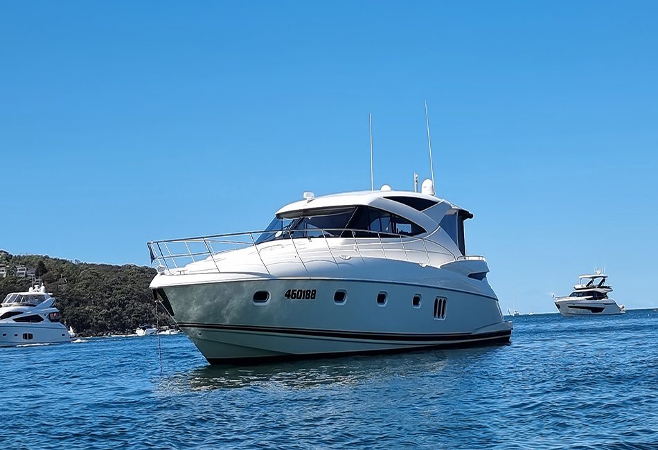 A white yacht with the registration number 460188 is anchored in calm blue waters. Other yachts are visible in the distance against a backdrop of a lush green shoreline and clear blue sky, offering a glimpse into the luxurious experiences provided by The Yacht Social Club Sydney Boat Hire.