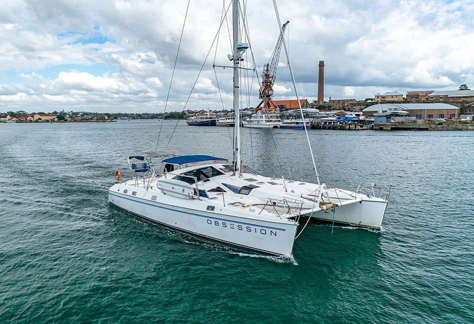 A white catamaran named "OBSESSION" sails on a calm body of water. The boat features blue-tinted windows and a blue bimini top. In the background, there is an industrial waterfront with cranes and buildings against cloudy skies. Experience this with Sydney Harbour Boat Hire from The Yacht Social Club.