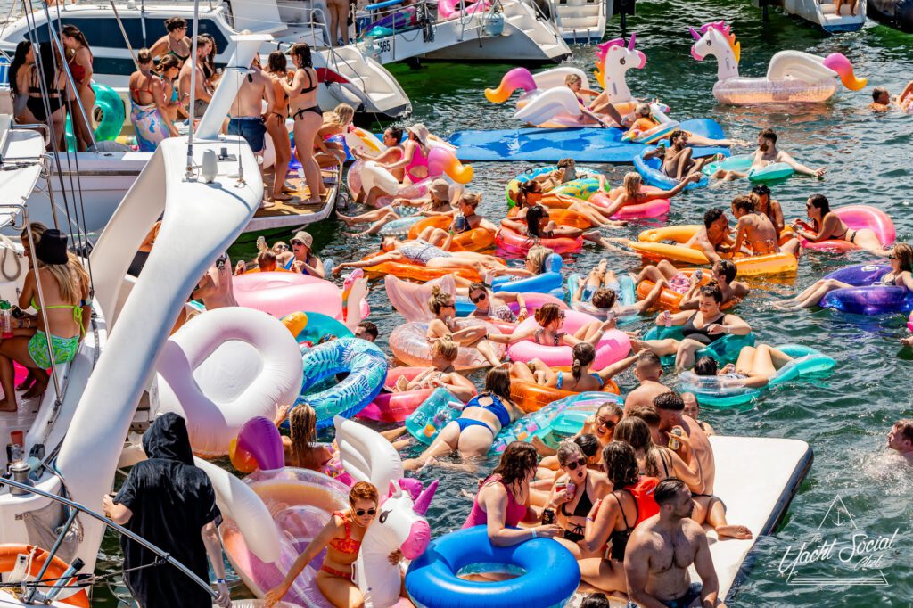 People are gathered for a lively pool party on inflatables of various shapes, including unicorns, donuts, and flamingos. The crowd is in swimsuits, enjoying the water and sunshine at The Yacht Social Club in Sydney, with luxury yacht rentals and boats docked in the background.