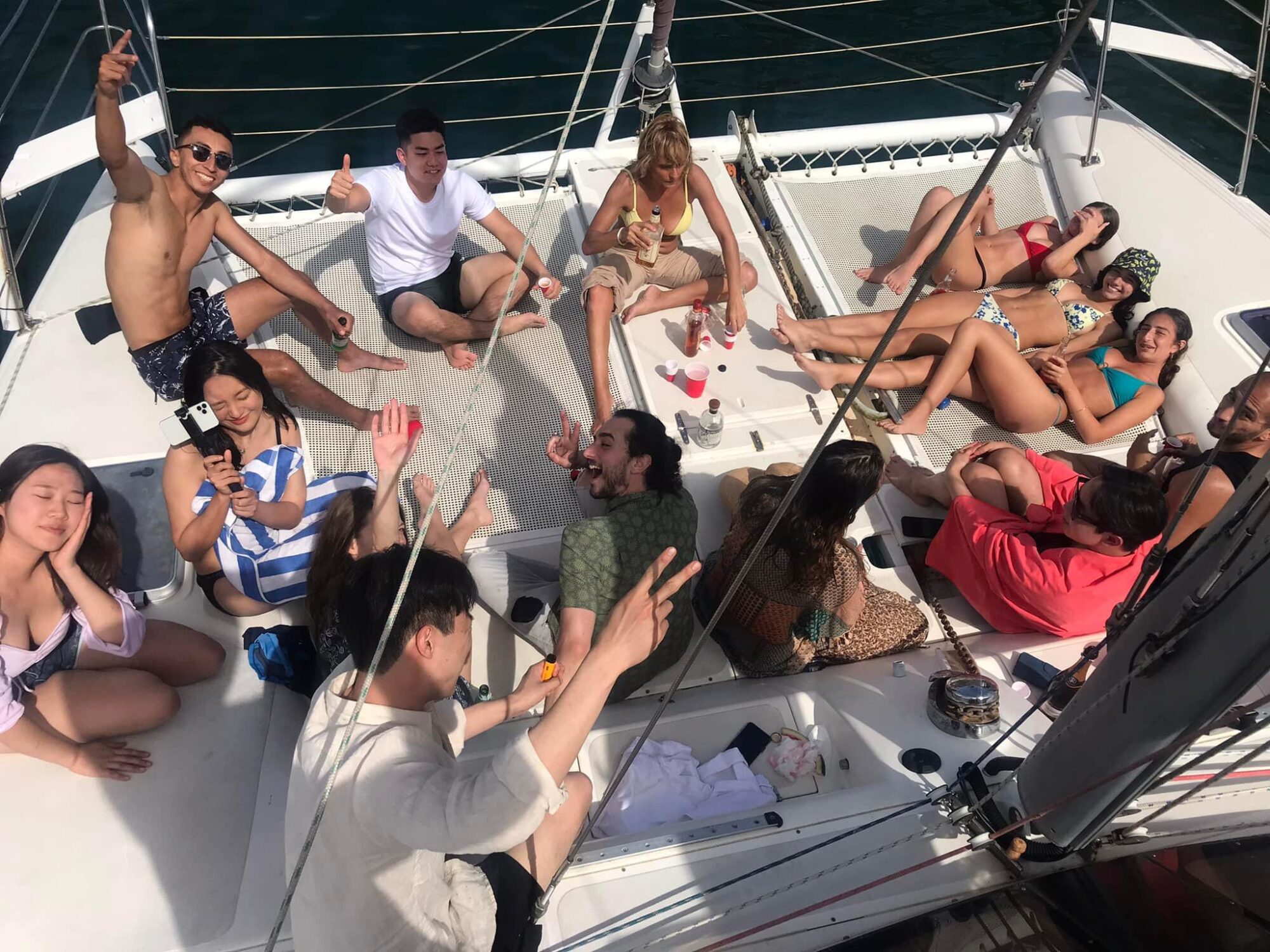 A group of people are gathered on the deck of a sailboat from The Yacht Social Club Sydney Boat Hire, enjoying themselves. Some are lounging on towels, while others are sitting or standing with smiles and raised hands. The day appears sunny, and everyone seems relaxed and cheerful.