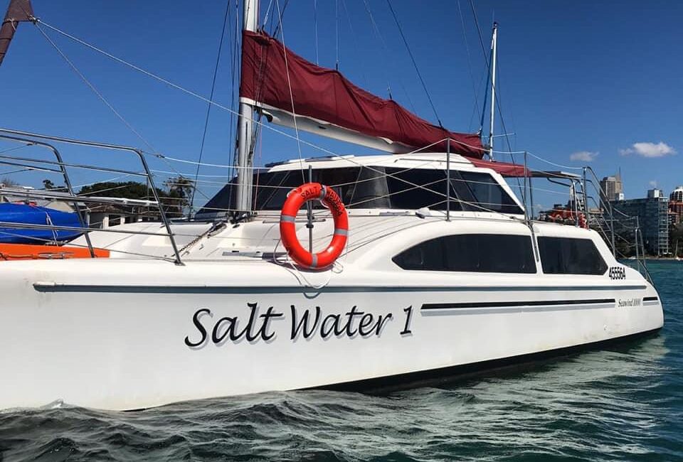 A white sailboat named "Salt Water 1" with a prominent life ring on its side is docked on clear, turquoise water. The boat, available through The Yacht Social Club, has a large red canopy and is set against a backdrop of blue sky and distant buildings.