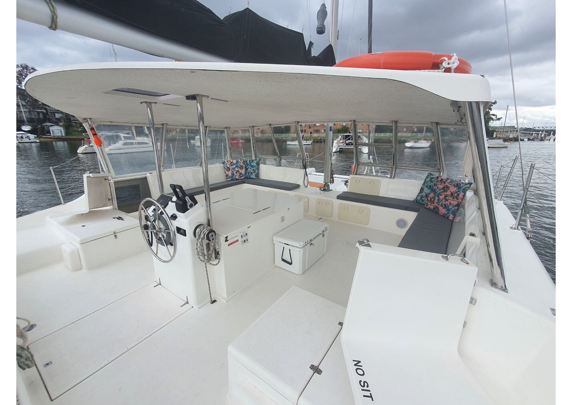 View of a boat's spacious outdoor seating area with a white canopy, gray cushions, and colorful throw pillows. The area includes a steering wheel, cooler, and ample seating. Perfect for Boat Parties with The Yacht Social Club in Sydney Harbour. The boat is docked near a marina with other boats visible in the background.