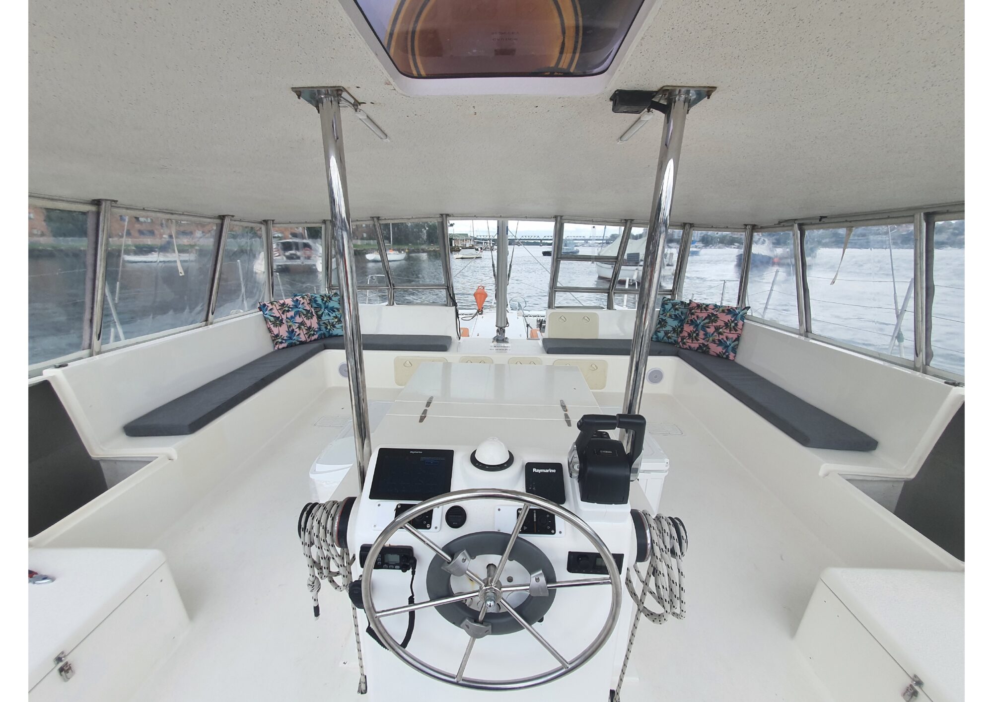 Interior view of a boat's cockpit with a steering wheel, navigational equipment, and various controls. The area features cushioned seating with colorful pillows around the sides and windows providing a view of the water and boats outside. Perfect for The Yacht Social Club Event Boat Charters or Luxury Yacht Rentals Sydney.