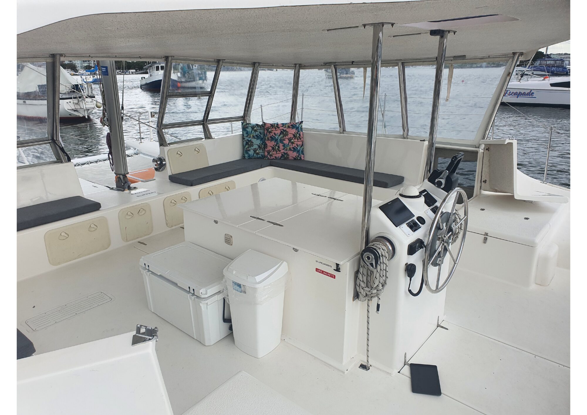 A covered outdoor seating area on a boat, featuring cushioned benches along the sides. A steering wheel and control panel are visible on the right. The area also includes a cooler and trash can. Beyond, other boats are seen docked on the water. Perfect for parties and events with The Yacht Social Club Sydney Boat Hire.