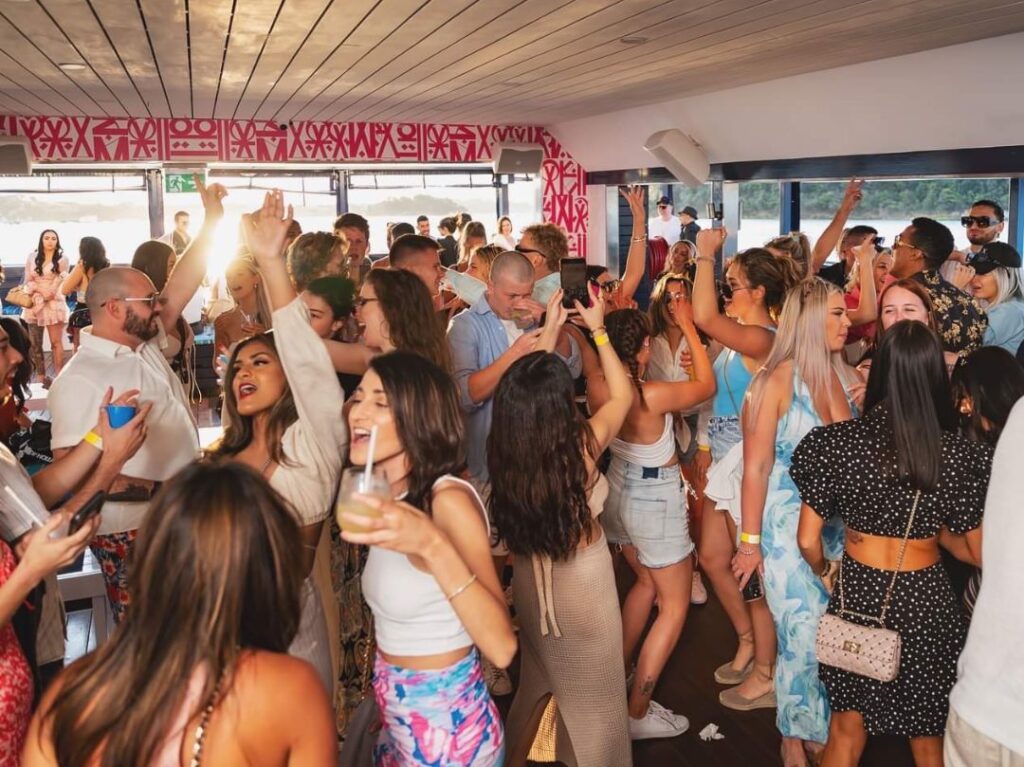 A lively indoor party with a large group of people dancing, talking, and enjoying drinks. The room is decorated with vibrant artwork on the walls. Some attendees are raising their hands and appear to be singing along to music. The atmosphere is energetic and festive, reminiscent of an event by Sydney Boat Hire's Yacht Social Club.