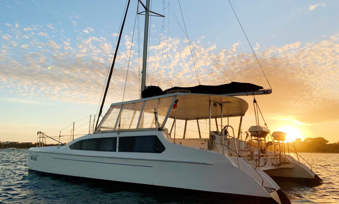 A white catamaran with multiple sails is anchored on calm waters during sunset. The sky is adorned with scattered clouds, and the sun casts a warm golden light on the scene, reflecting off the water. Trees are visible on the distant shore. Perfect for The Yacht Social Club Event Boat Charters or private gatherings!