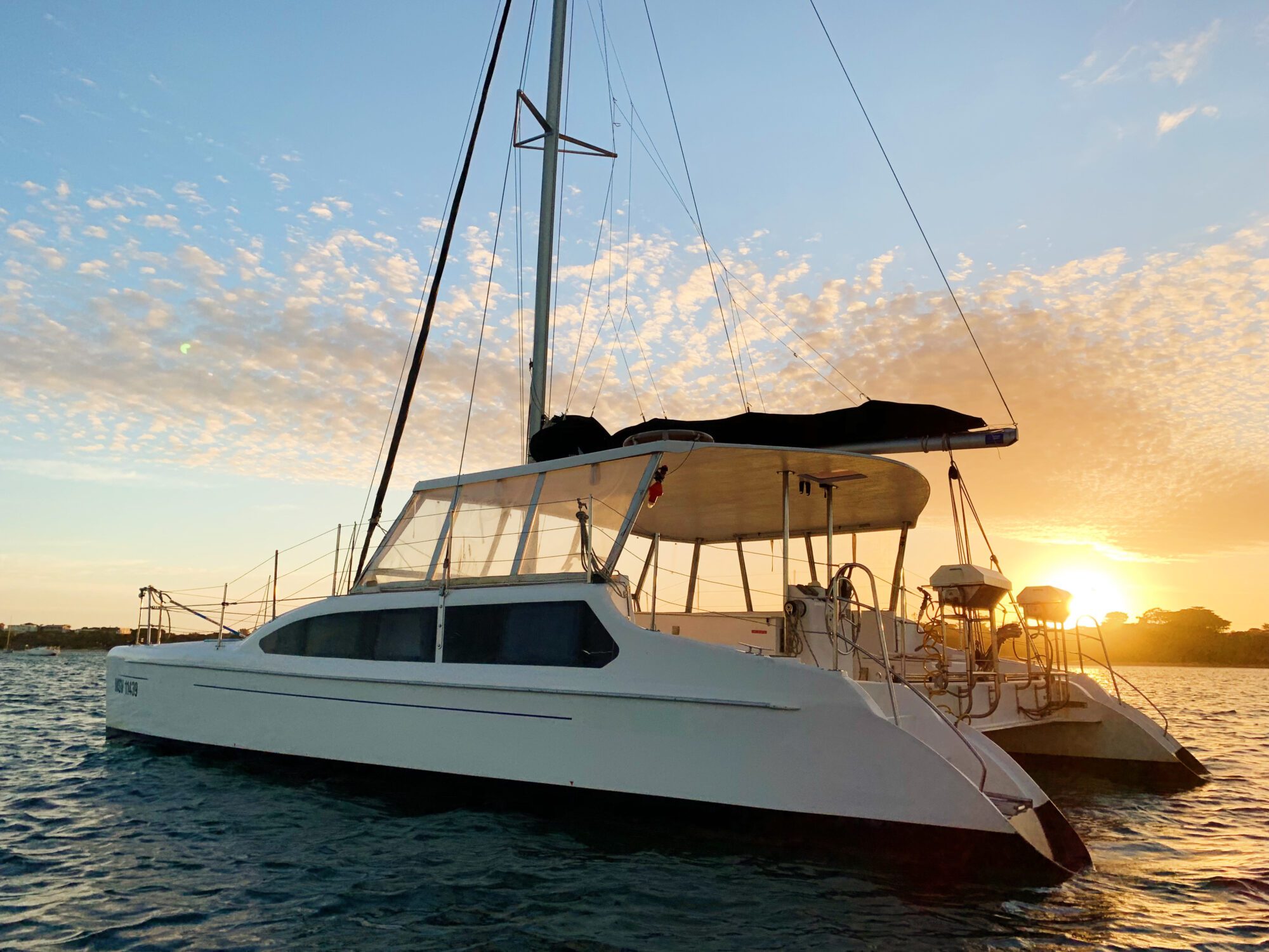 A white catamaran with multiple sails is anchored on calm waters during sunset. The sky is adorned with scattered clouds, and the sun casts a warm golden light on the scene, reflecting off the water. Trees are visible on the distant shore. Perfect for The Yacht Social Club Event Boat Charters or private gatherings!