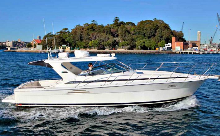 A white motorboat with a sleek design glides through blue water. A person is piloting the boat, which features a spacious deck and several railings. In the background, an island with dense trees and some buildings is visible under a clear sky. Experience this with Sydney Harbour Boat Hire The Yacht Social Club.